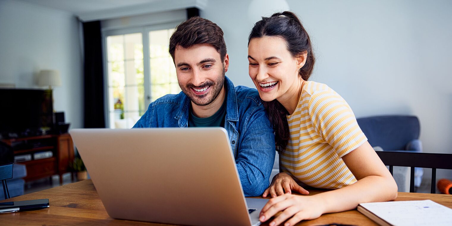 Frau und Mann sitzen an einem Tisch und blicken in einen Laptop. Junge Frau und junger Mann sitzen zusammen an einem Tisch und blicken mit Freude in einen Laptop.
