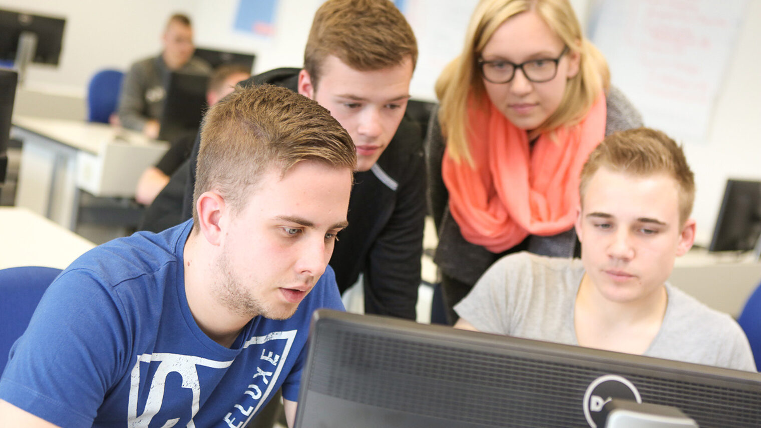 Kursteilnehmer sitzen vor einem Computer. Eine Kursteilnehmerin blickt ihnen dabei über die Schulter.