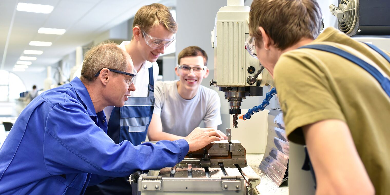 Ein Meister erklärt in der Werkstatt drei Auszubildenden einen Arbeitsschritt.