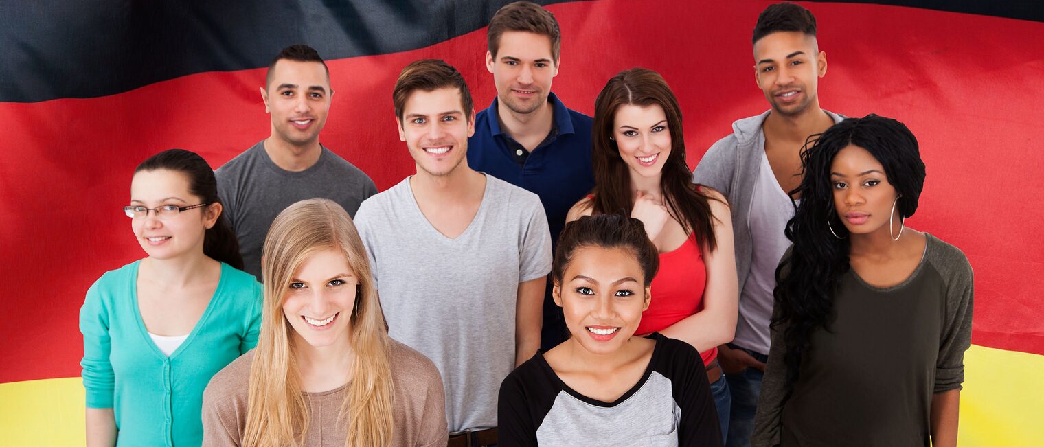 Junge Menschen mehrerer Nationalitäten stehen vor einer Deutschlandfahne.