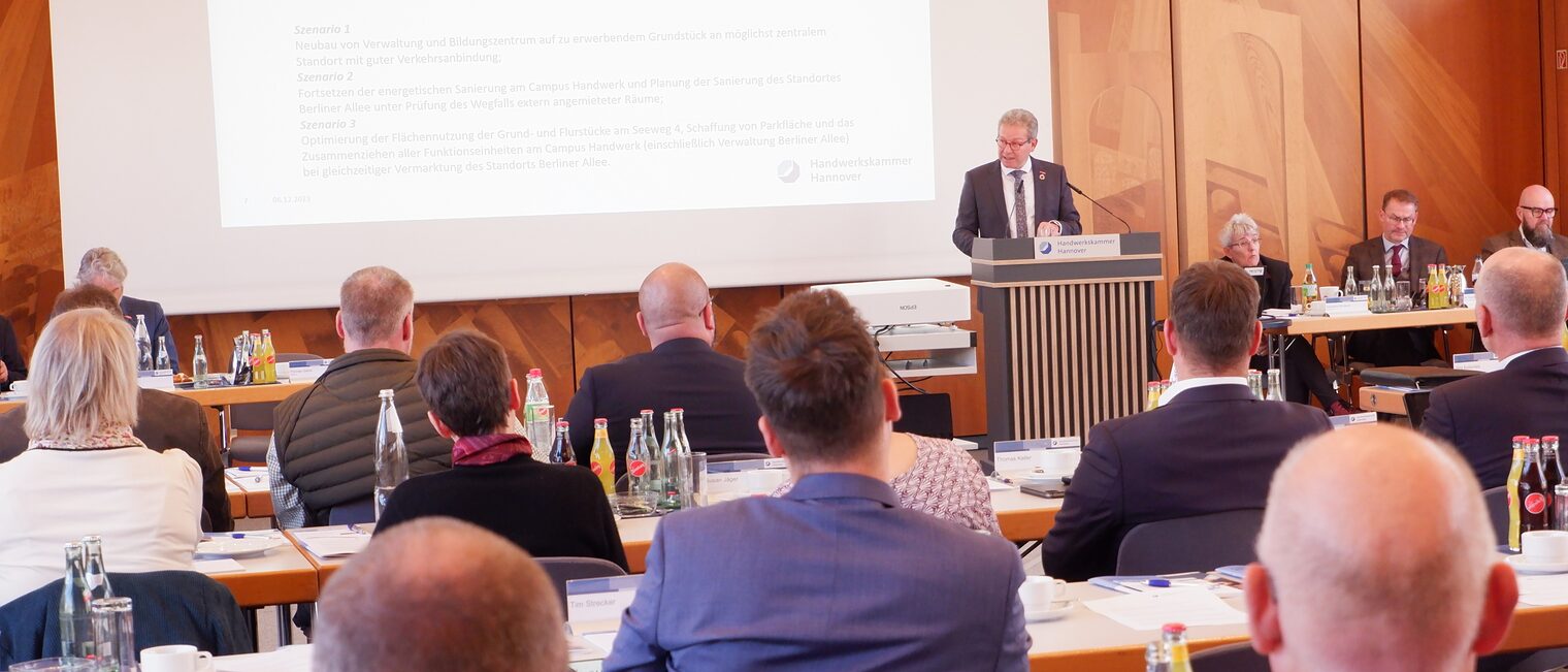 Mann am Pult spricht vor Menschen an Tischen.