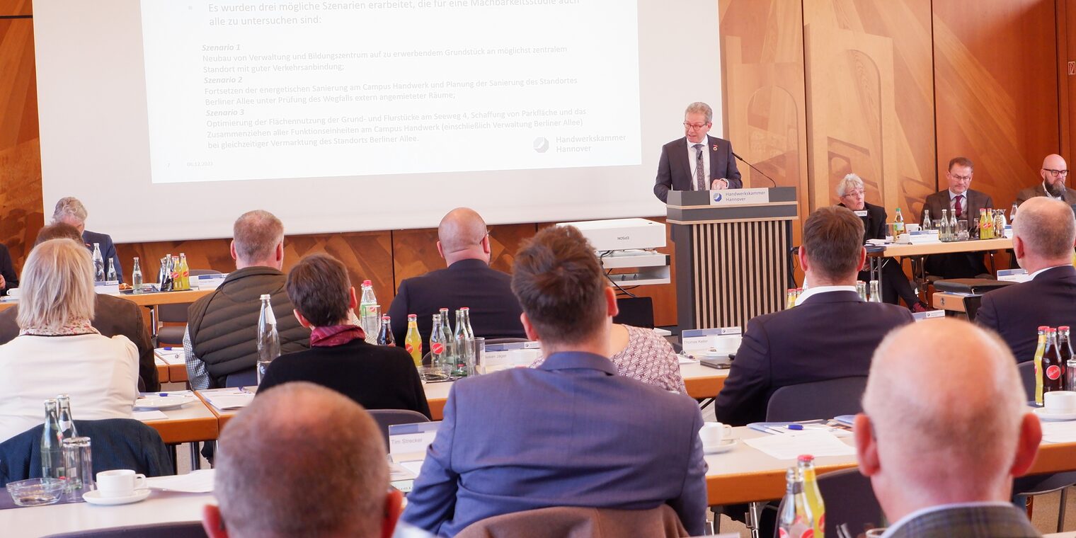 Mann am Pult spricht vor Menschen an Tischen.