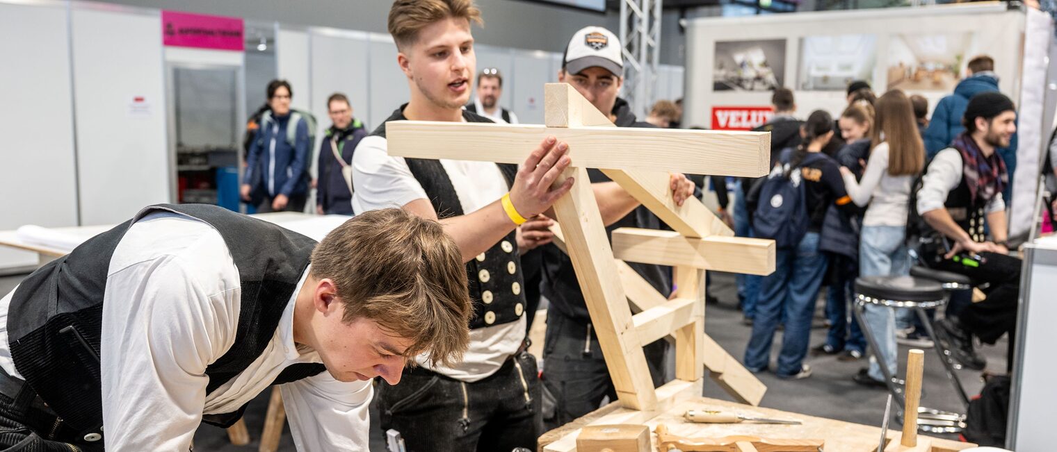 Zwei Zimmerer bauen an einem Objekt aus Holz bei der IHM in einer Messehalle.