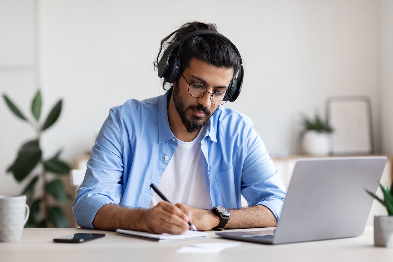 Junger Mann sitzt mit einem Laptop und Kopfhörer am Tisch und notiert sich etwas auf Papier.