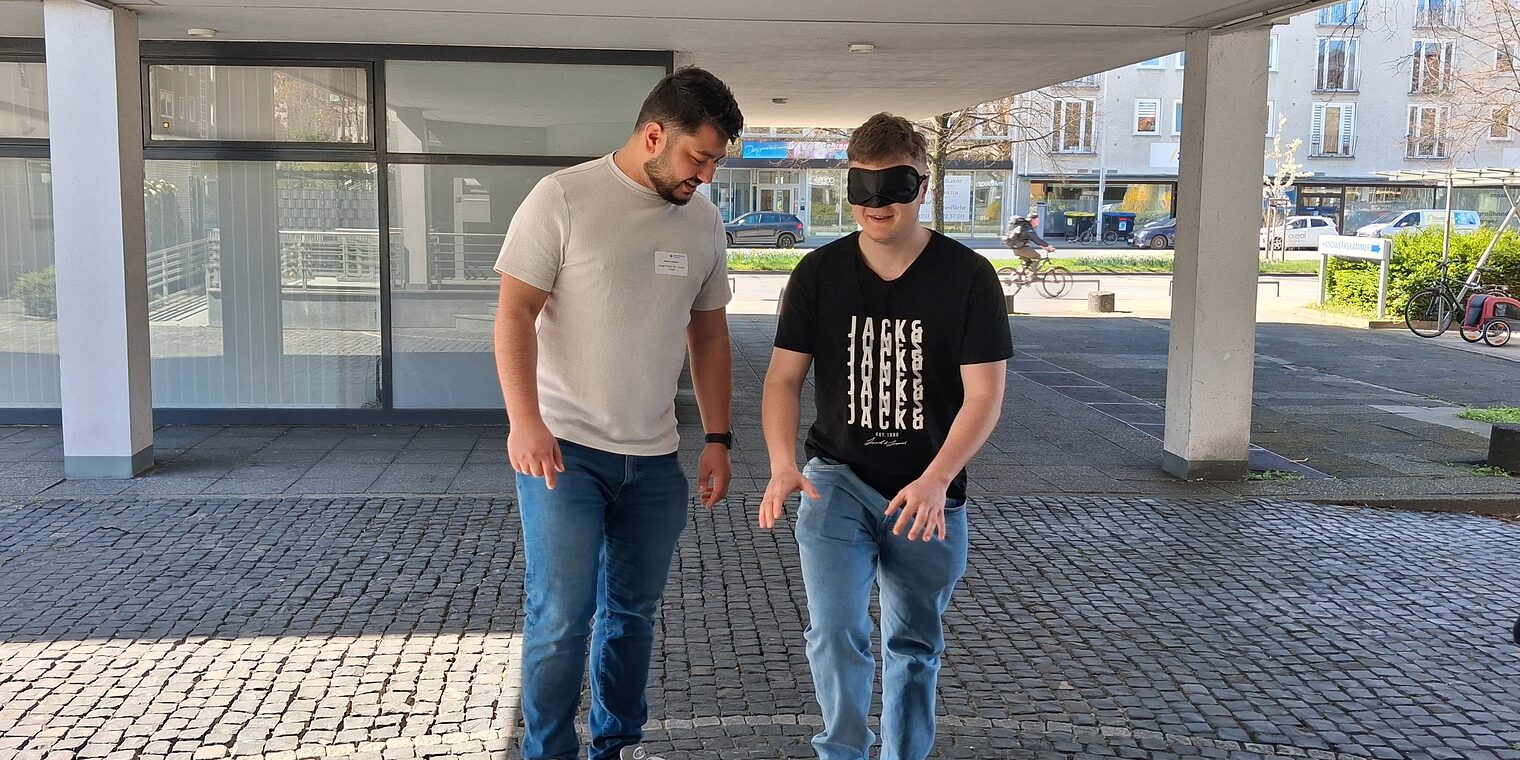 Ein Geselle lotst einen anderen mit verbundenen Augen drei Treppenstufen hoch.