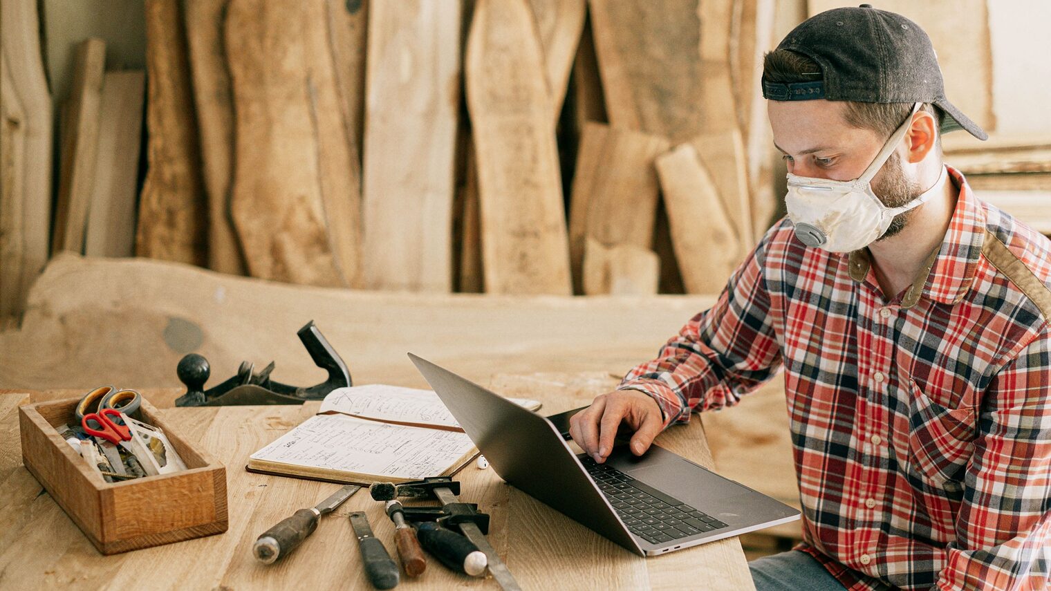Handwerker sitzt mit FFP-Maske an seinem Laptop in der Werkstatt. Auf dem Tisch liegen Werkzeuge und ein Notizbuch.