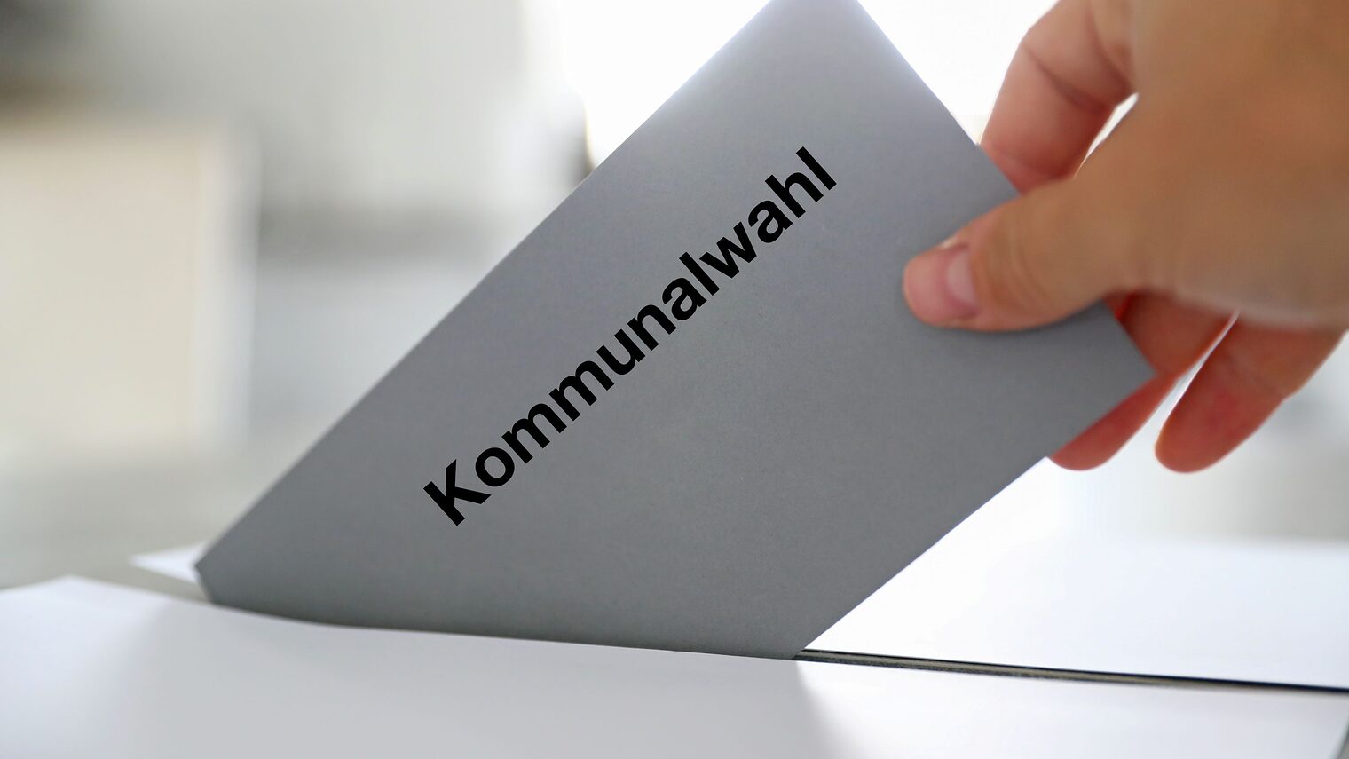Hand steckt Wahlzettel mit Kommunalwahl-Aufschrift in Wahlurne.