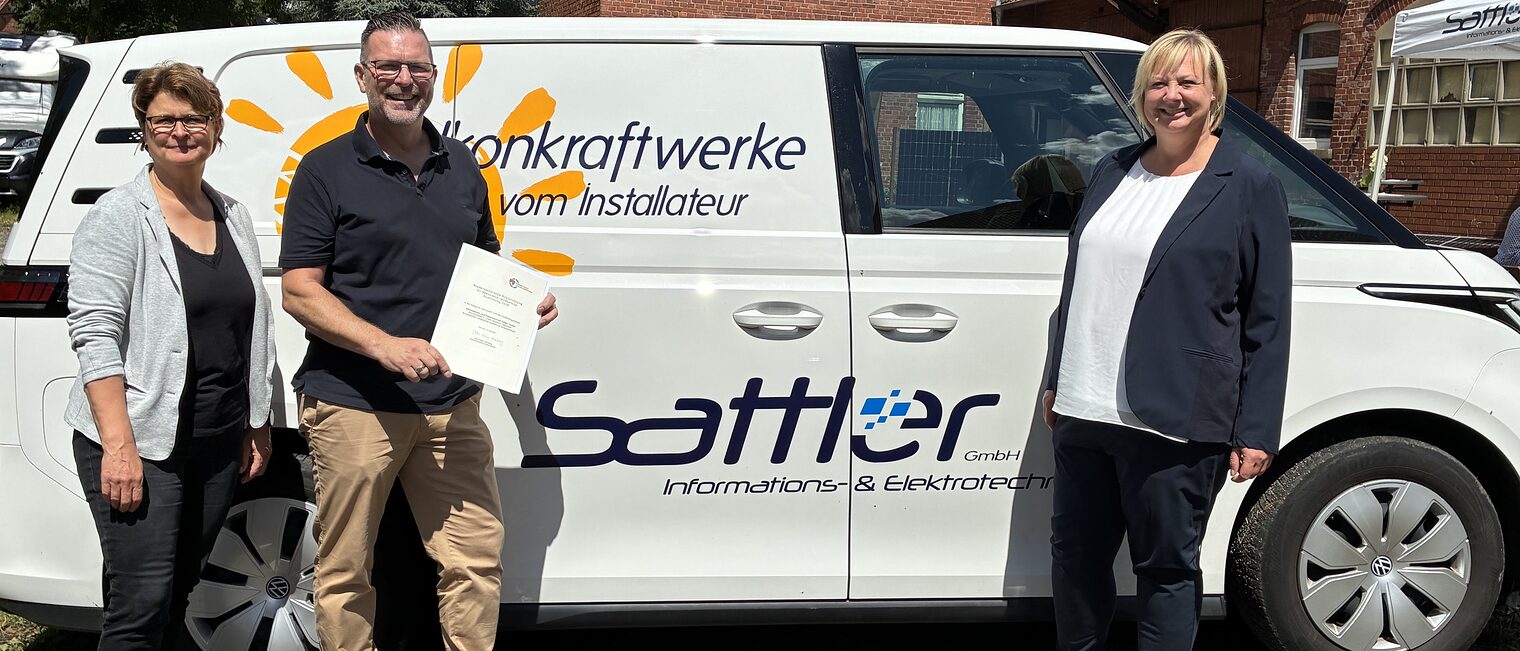 Geschäftsführer Sven Sattler, Ute Sandtvos (Vertreterin des Kultusministeriums) und Bettina Wolf-Moritz (HWK Hannover) bei der Verleihung des Preises für besonders verlässliche Ausbildung vor dem Firmenwagen. 